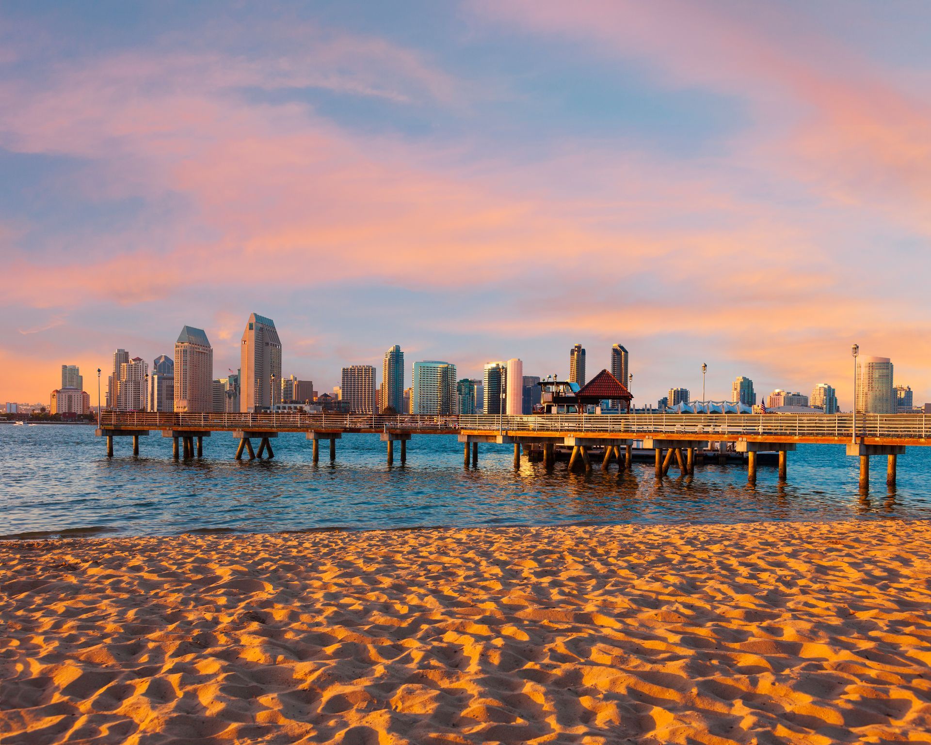 Beach with a pier, city skyline at sunset. Golden sand, pink/orange sky, blue water.