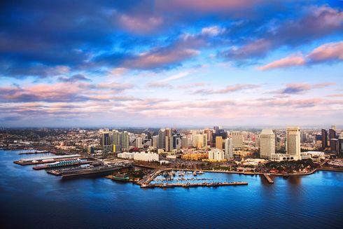 City skyline by a bay under a cloudy sky. Water in the foreground, buildings and boats in the middle.