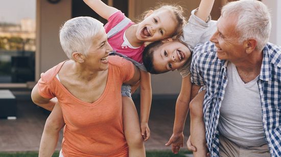Grandparents give piggyback rides to laughing grandchildren in a sunny yard.