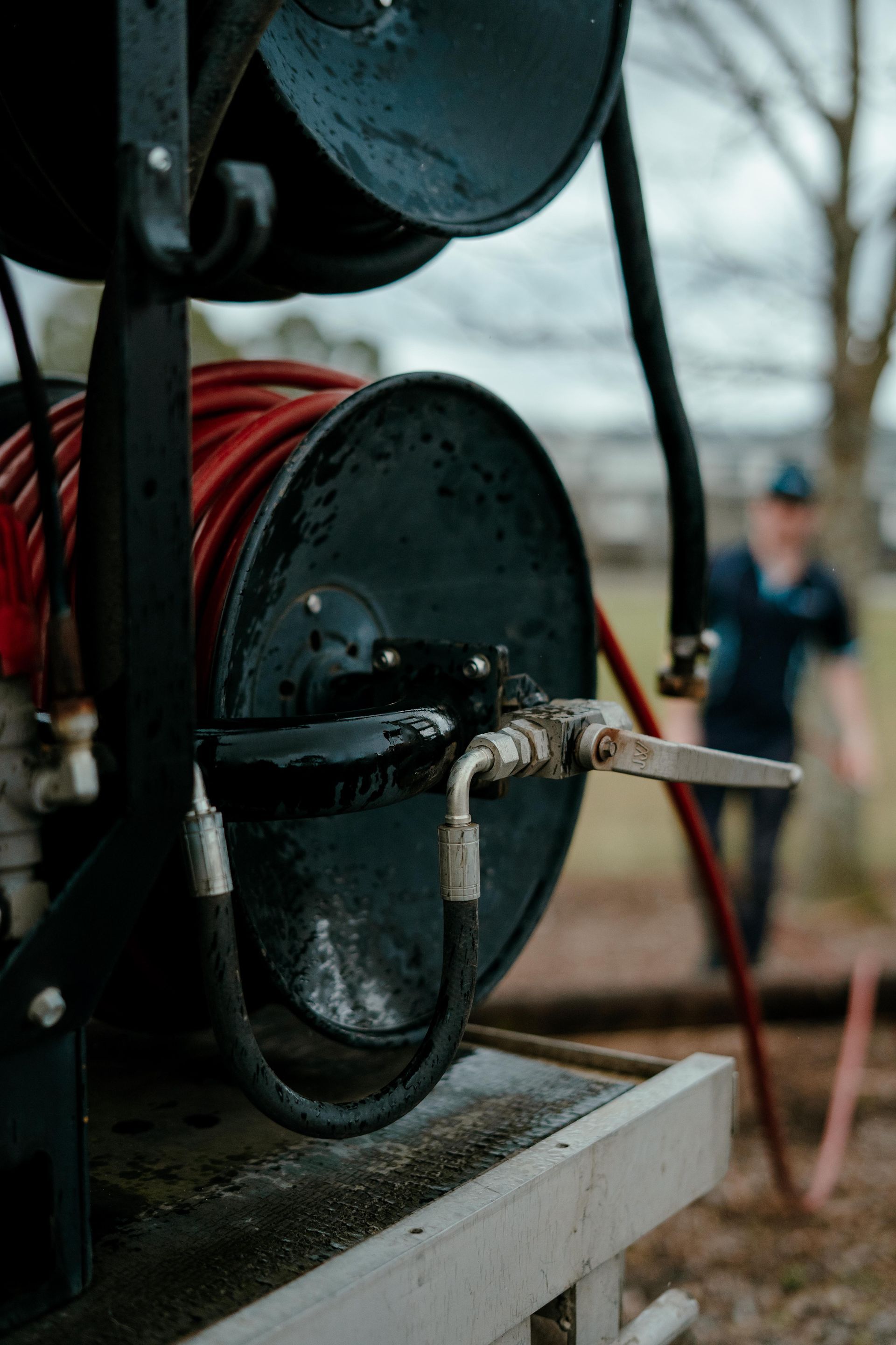 Close-up of a red cable reel and hose on a metal cart outdoors, with a blurred worker in the background — Aquafix Plumbing Services in Bowral, NSW