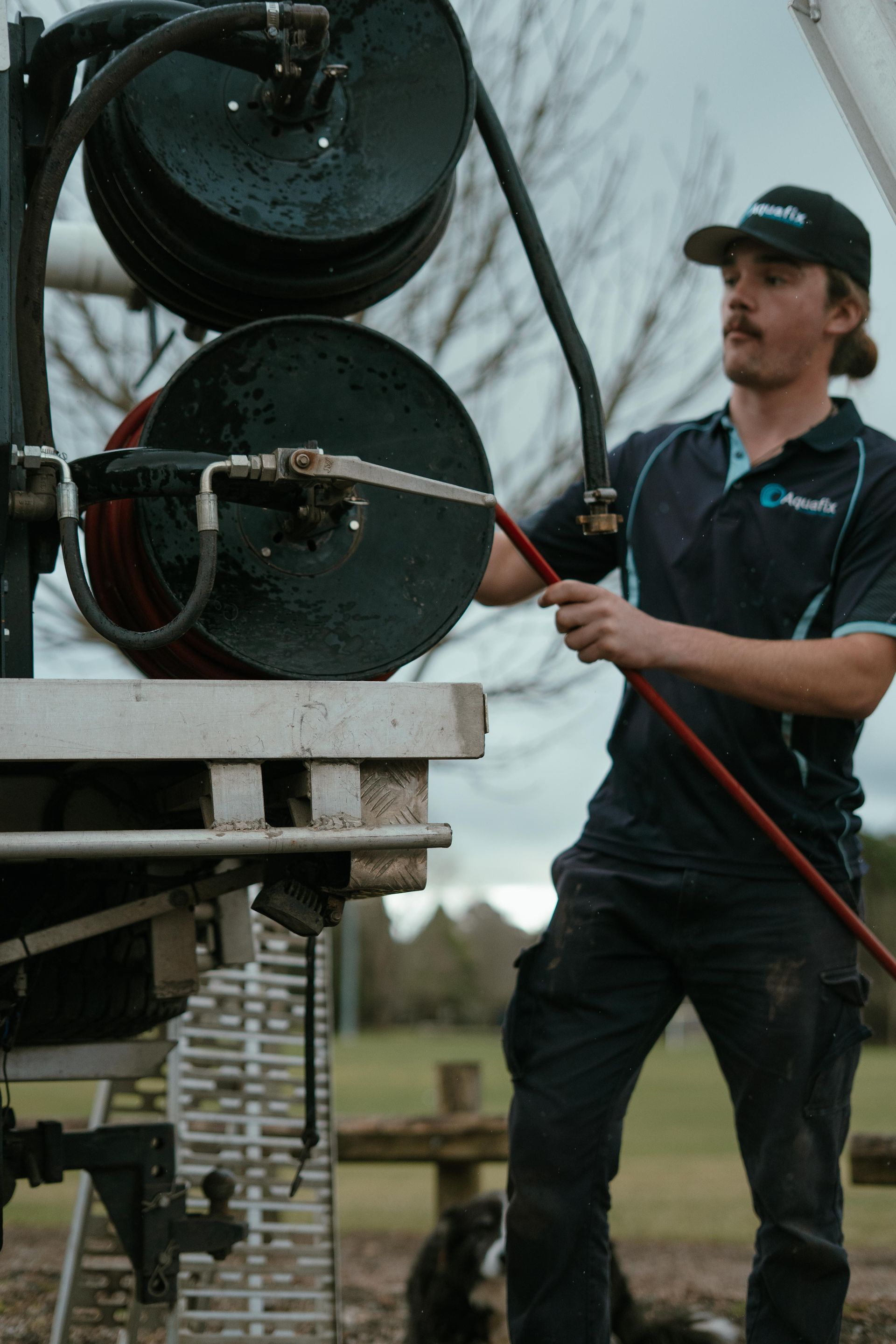Worker in black uniform handling a red hose beside stacked large black barrels outdoors — Aquafix Plumbing Services in Bowral, NSW