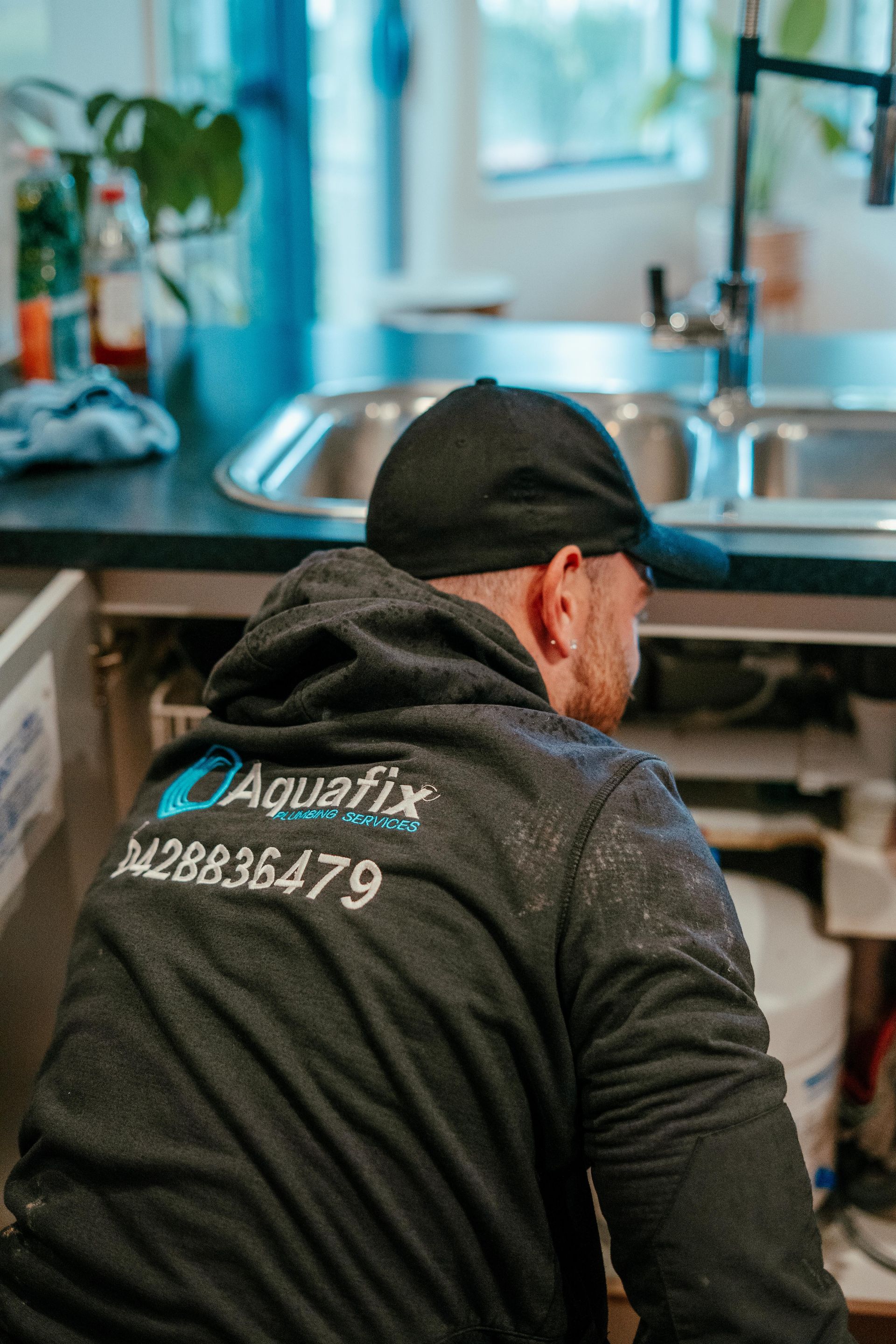 Worker in black jacket under a sink in a kitchen, repairing plumbing. — Aquafix Plumbing Services in Bowral, NSW