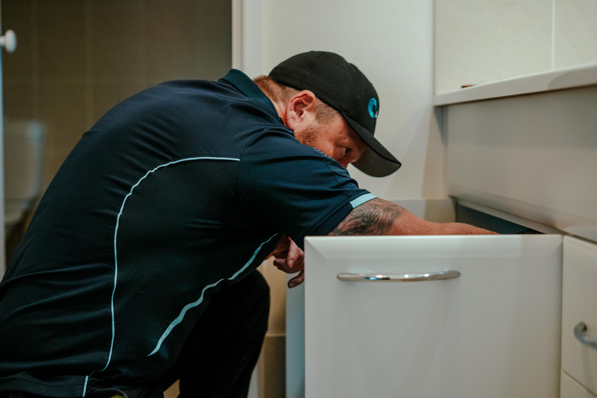 Person in black cap and jacket leaning into an open cabinet, working in a kitchen. — Aquafix Plumbing Services in Bowral, NSW