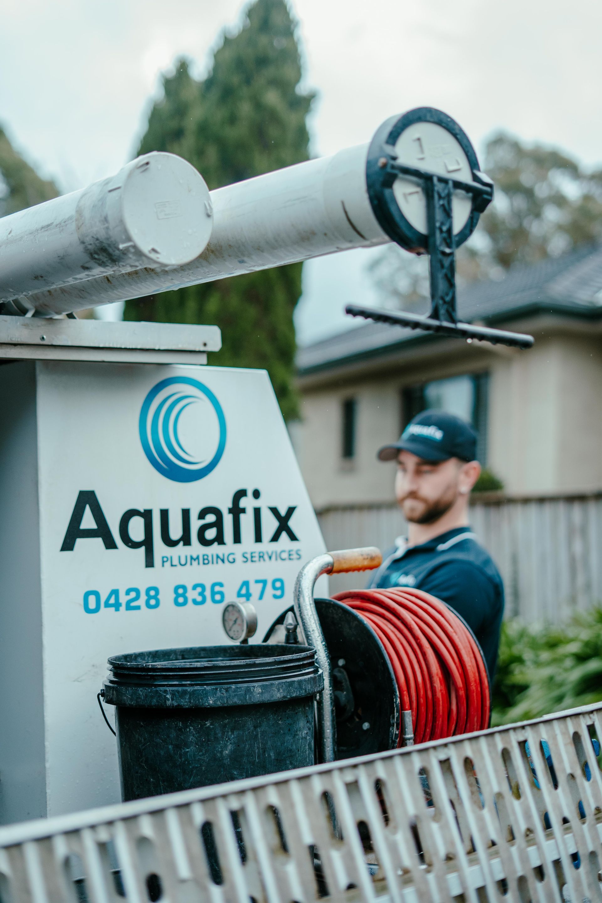 Aquafix service truck with pipes and hose reel, worker in background beside a fence — Aquafix Plumbing Services in Bowral, NSW