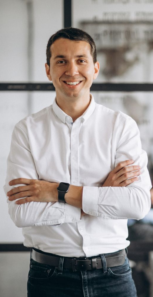 Man with arms crossed smiling, wearing a white shirt and watch. In an office setting.