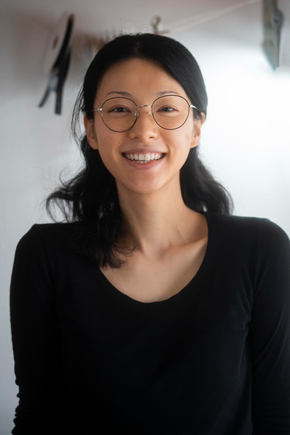 Woman with round glasses smiling, wearing a black shirt, in front of a white wall.