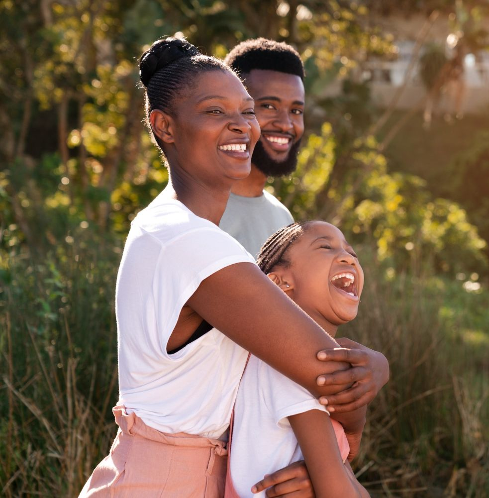 Family laughing outdoors: mother hugs daughter, father behind them smiles. Warm sunlight.