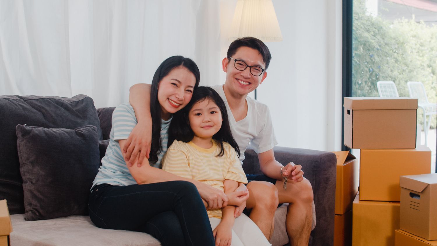 Family of three smiling, sitting on a sofa in a living room with moving boxes.