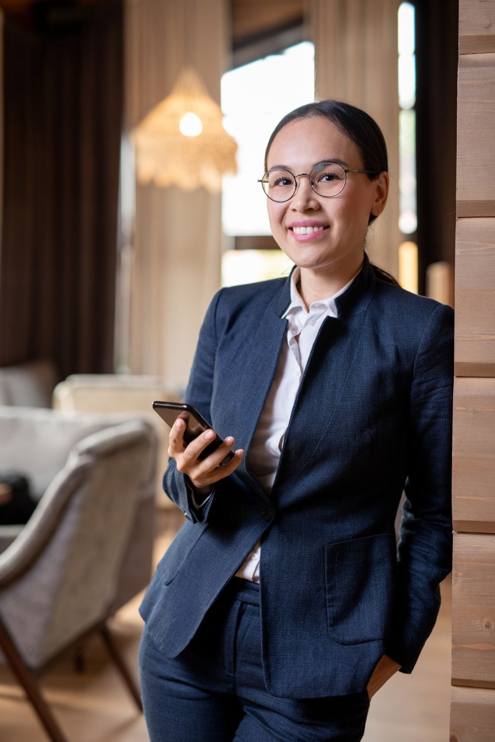 Woman in a navy suit and glasses holds a phone, leaning against a wooden wall, smiling.