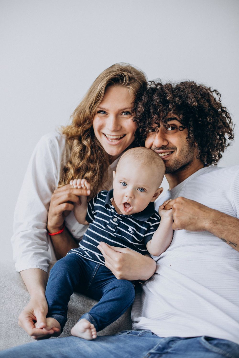 Family of three poses indoors. A baby is held by parents, smiling at the camera.