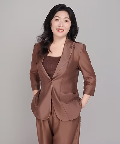 Woman in a brown pantsuit smiling, hands in pockets, against a gray background.