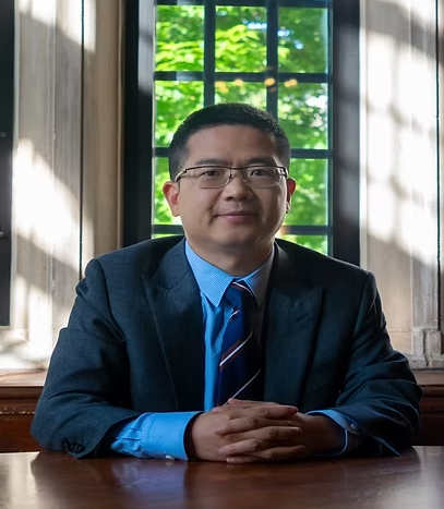 Man in suit, seated at desk, hands clasped, in front of a window with greenery.