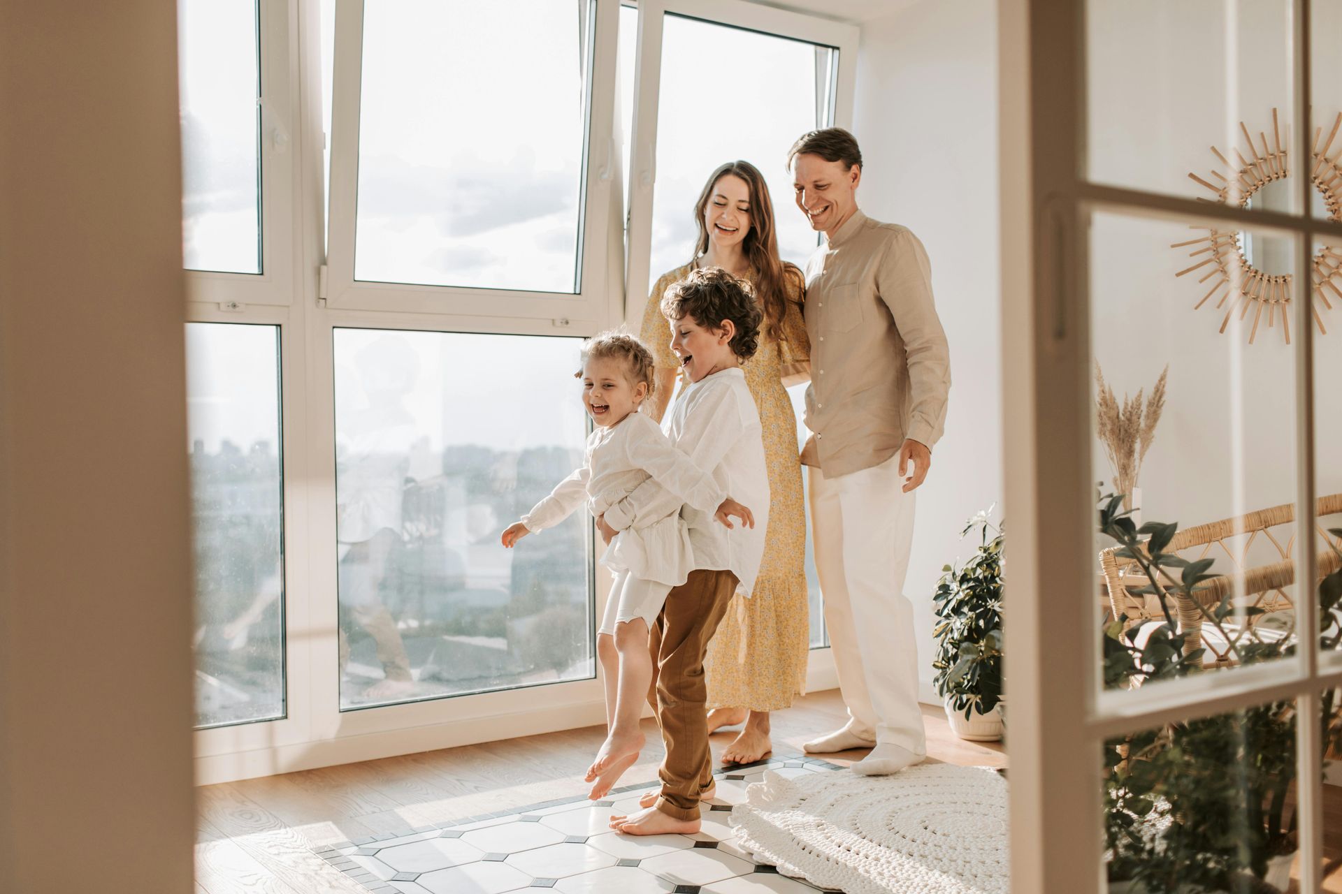 Family of four standing by a large window, laughing. Sunlit room, neutral tones, happy expressions.
