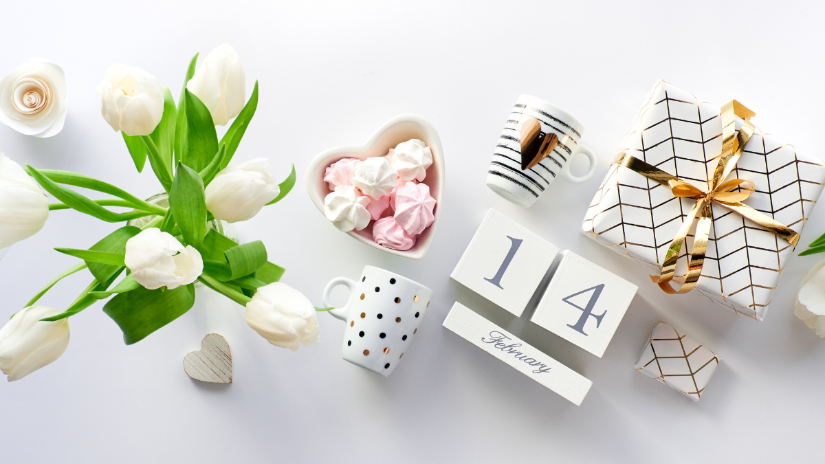 White tulips, heart-shaped dish of meringues, mugs, wrapped gift, and Valentine's Day calendar on white.