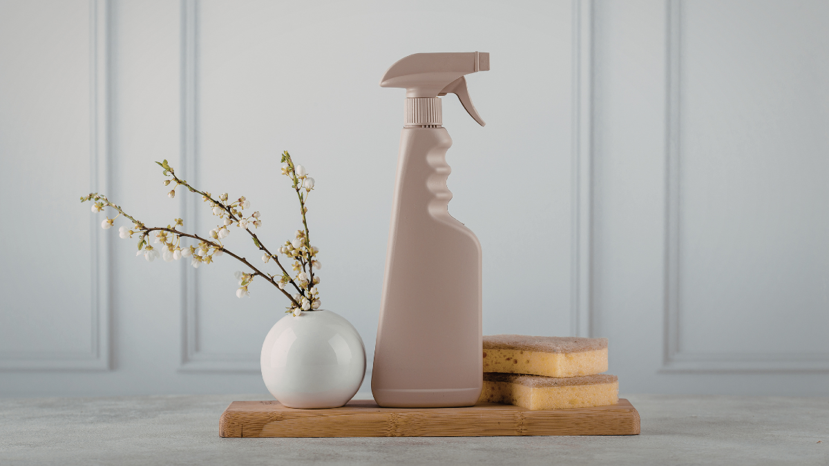 A beige spray bottle, two yellow sponges, and a small white vase with sprigs of white flowers on a wooden board.