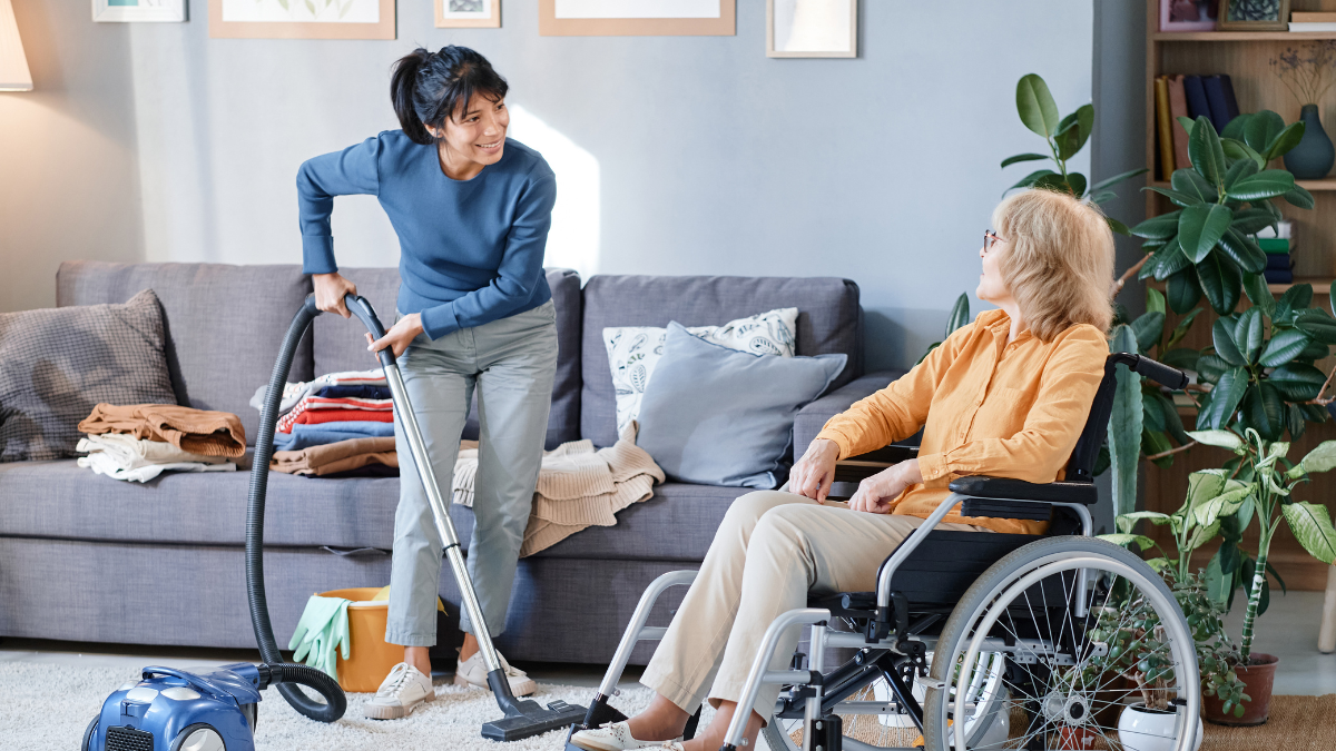 A woman is vacuuming the floor next to an elderly woman in a wheelchair.