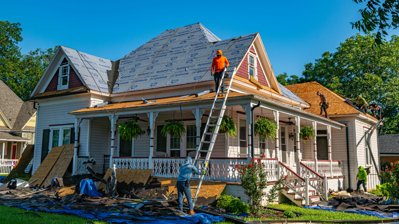 Two roofers replacing shingles on a house roof; ladder, blue tarp, trees in background.