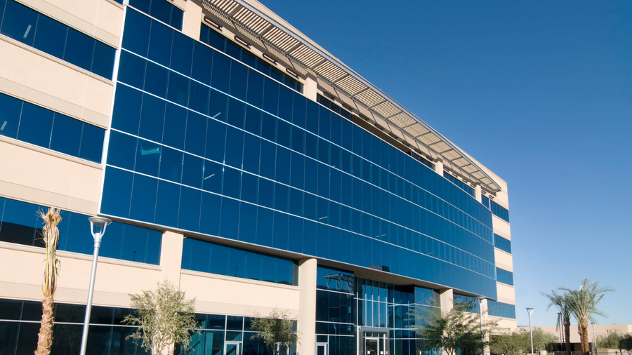 Modern office building with blue-tinted windows reflecting the sky, under a bright, sunny day.