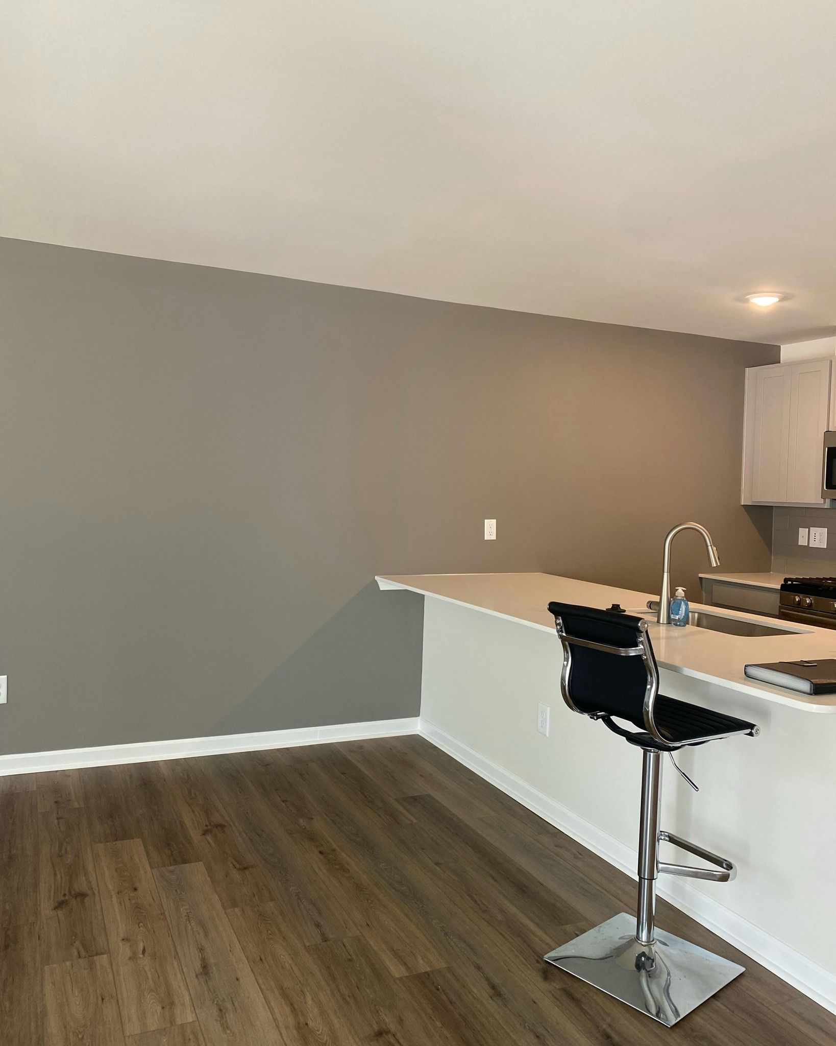 Kitchen with gray wall, white countertop, and a black bar stool. Brown wood-look floor.
