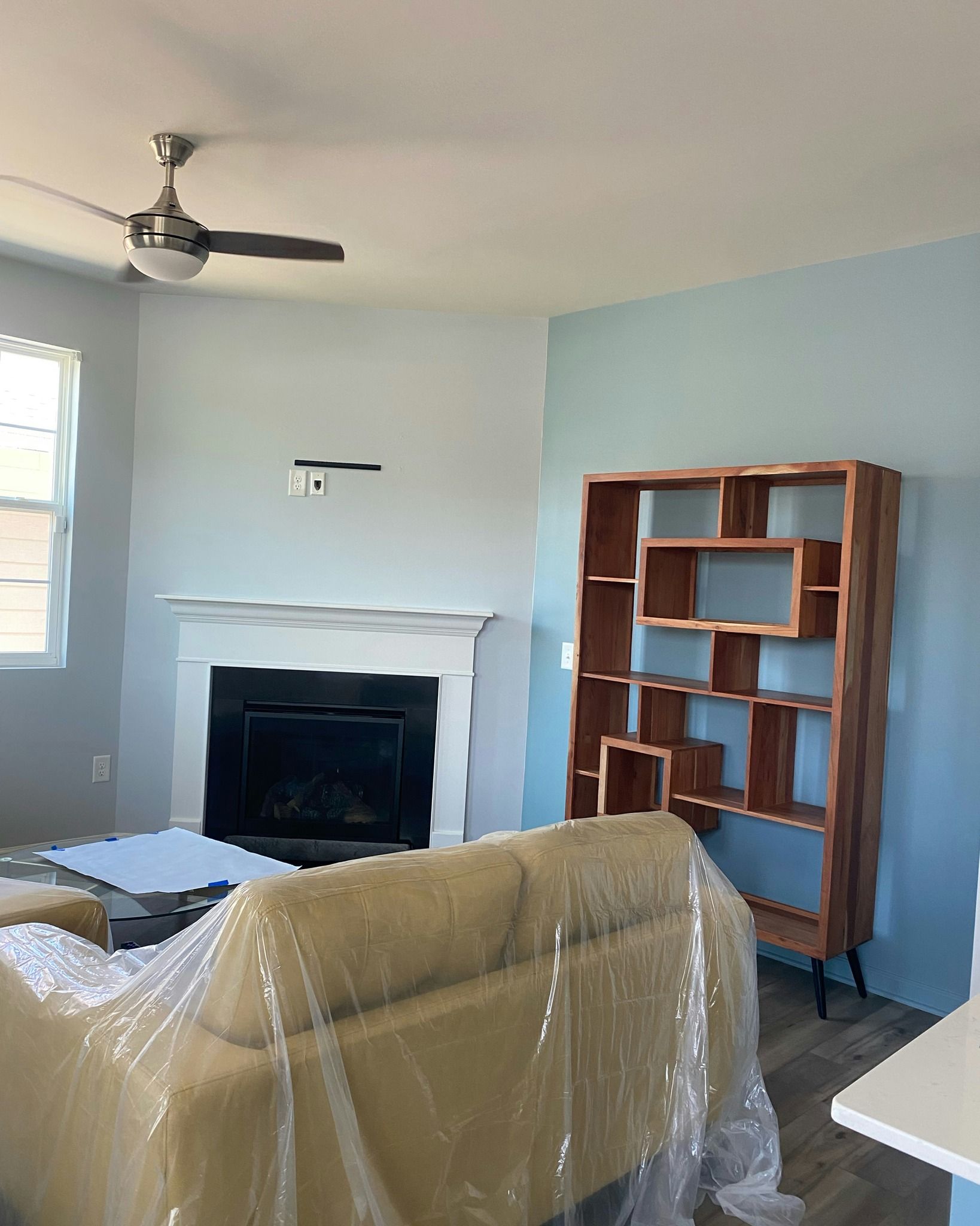 Living room with fireplace, bookshelf, and sofa covered in plastic. Pale blue walls and ceiling fan.