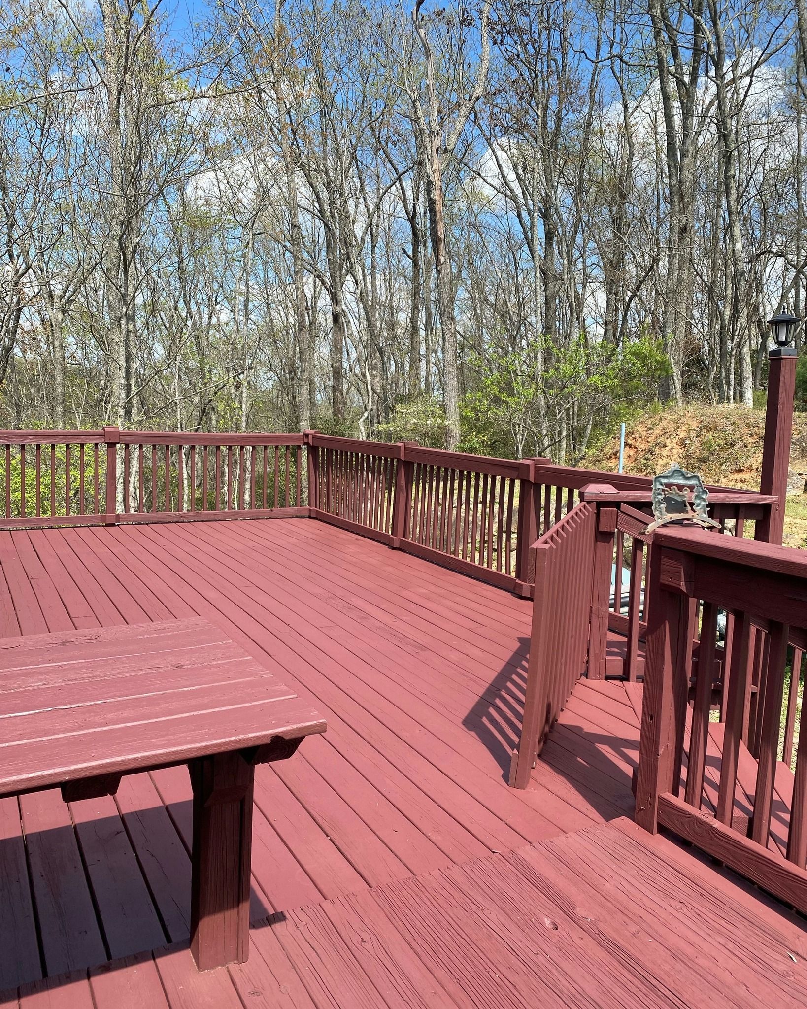 Red wooden deck in a wooded area with a table, railing, and a sunny blue sky.