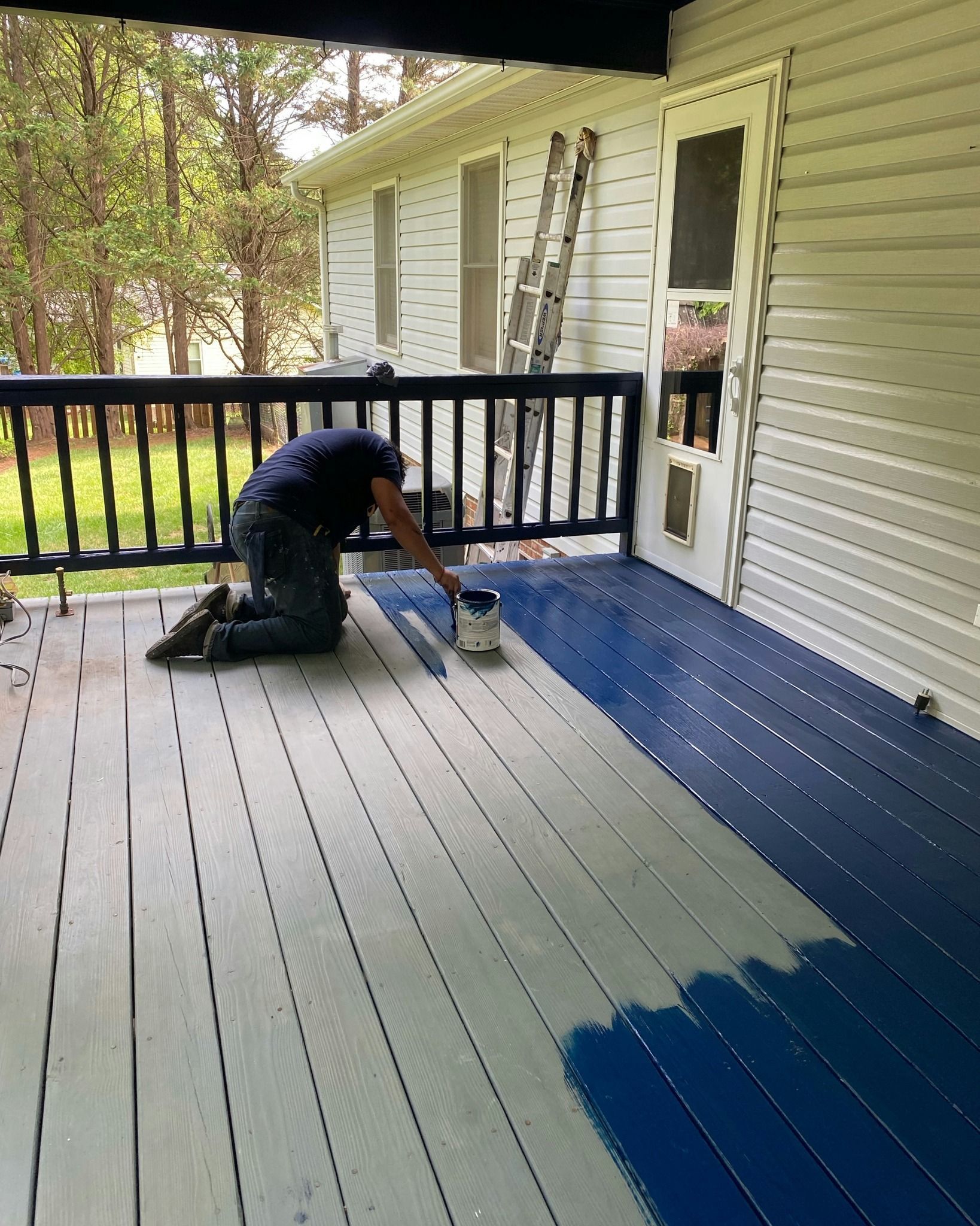 Person kneeling on a deck, painting it blue.  Half the deck is painted blue, the other half is unpainted.