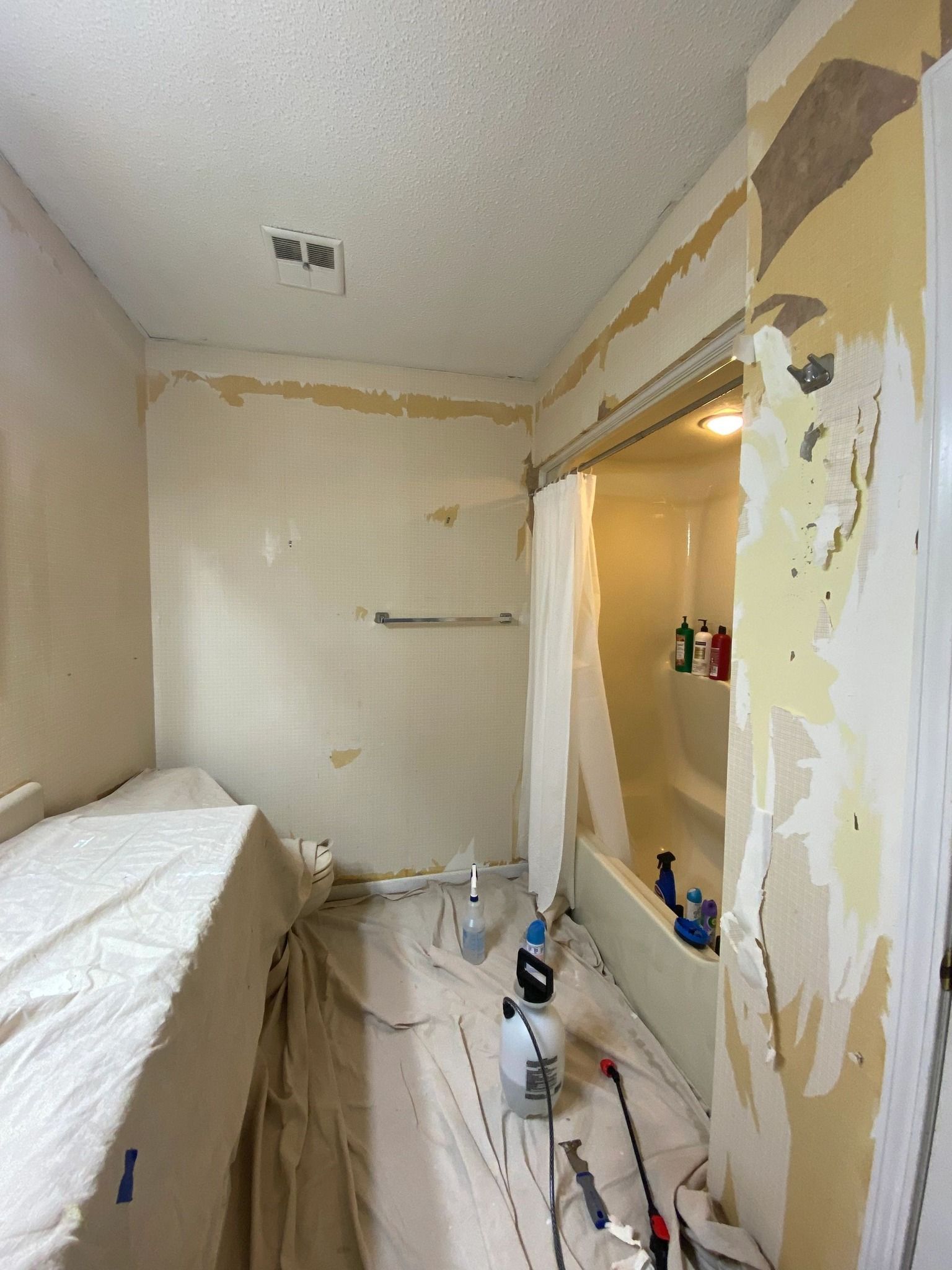 Bathroom being renovated: walls stripped of wallpaper, covered with drop cloths, and tools visible.