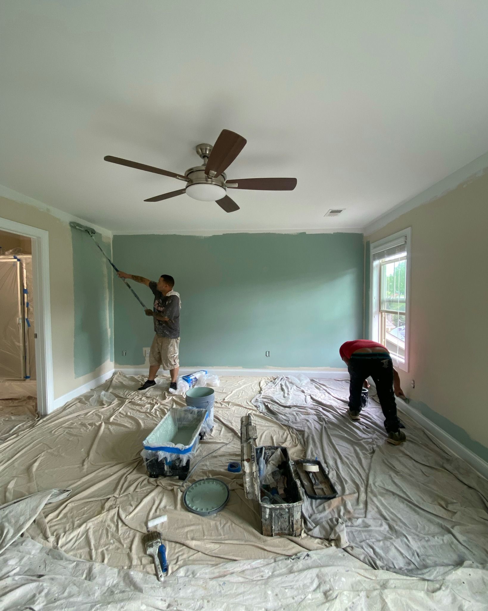 Two painters painting a room with light green and beige walls, drop cloths on the floor, a ceiling fan.