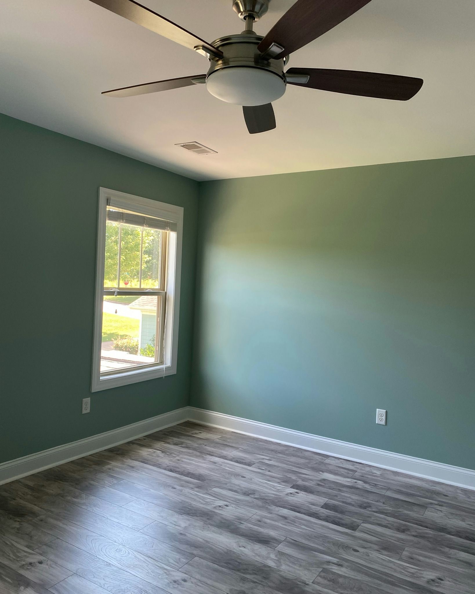 Empty bedroom with teal walls, a ceiling fan, window, and gray wood-look flooring.