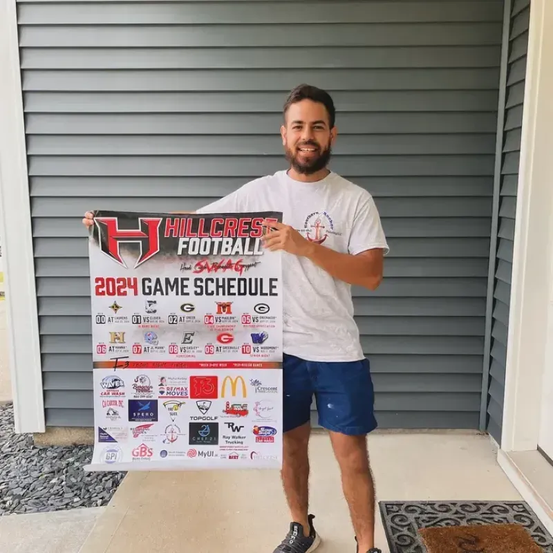 Man holding a 2024 Hillcrest Football game schedule poster. He's smiling outside a grey-sided house.