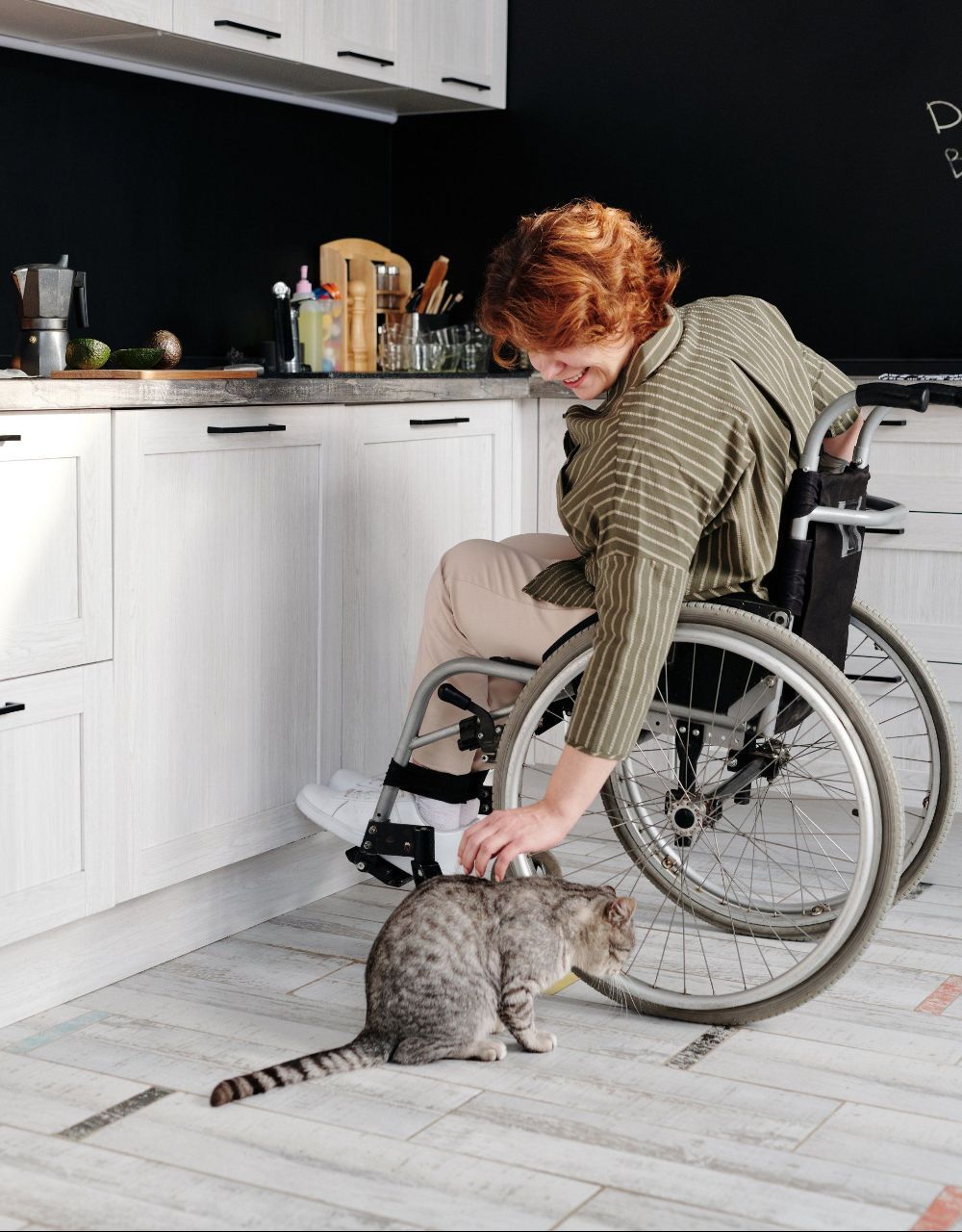 A woman in a wheelchair is petting a cat in a kitchen.