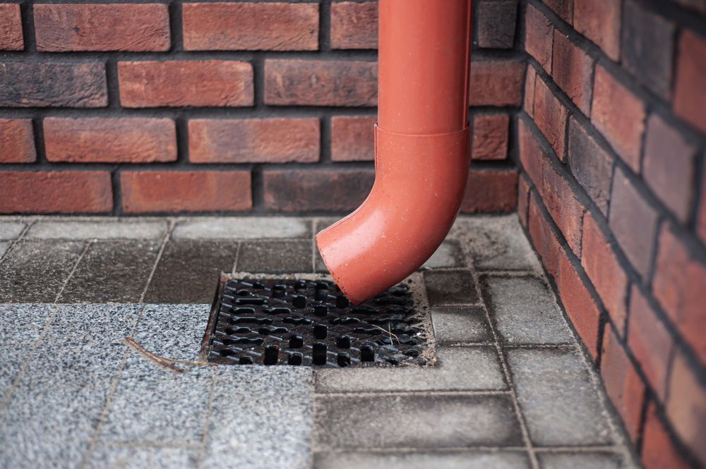 A red pipe is coming out of a drain on the sidewalk next to a brick wall.