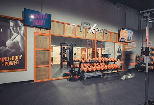 Gym interior: orange accents, weights, TV screens, woman on a poster, person exercising.