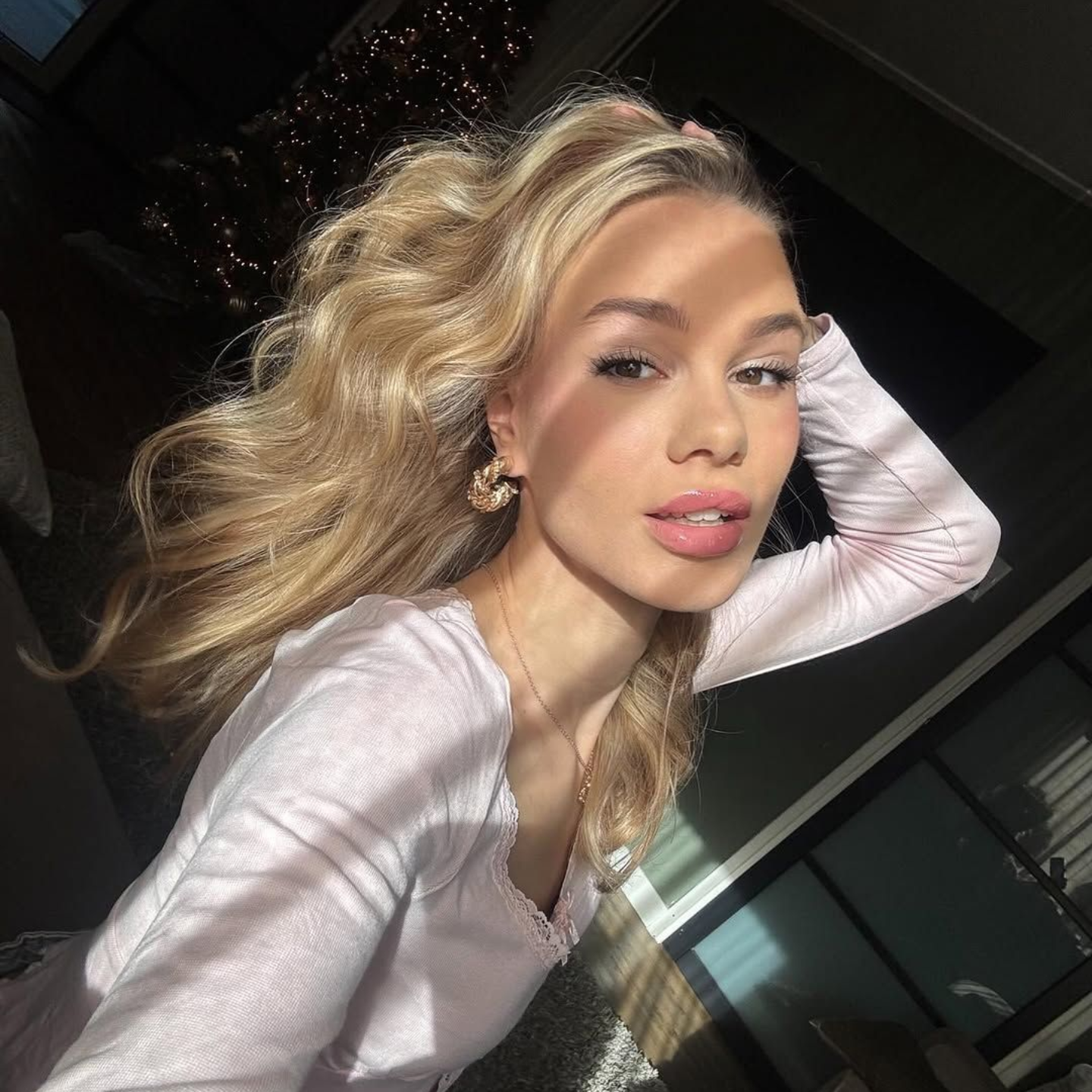 Een persoon met golvend blond haar draagt ​​een lichtgekleurde top en poseert met een hand achter het hoofd in een zonovergoten binnenruimte.