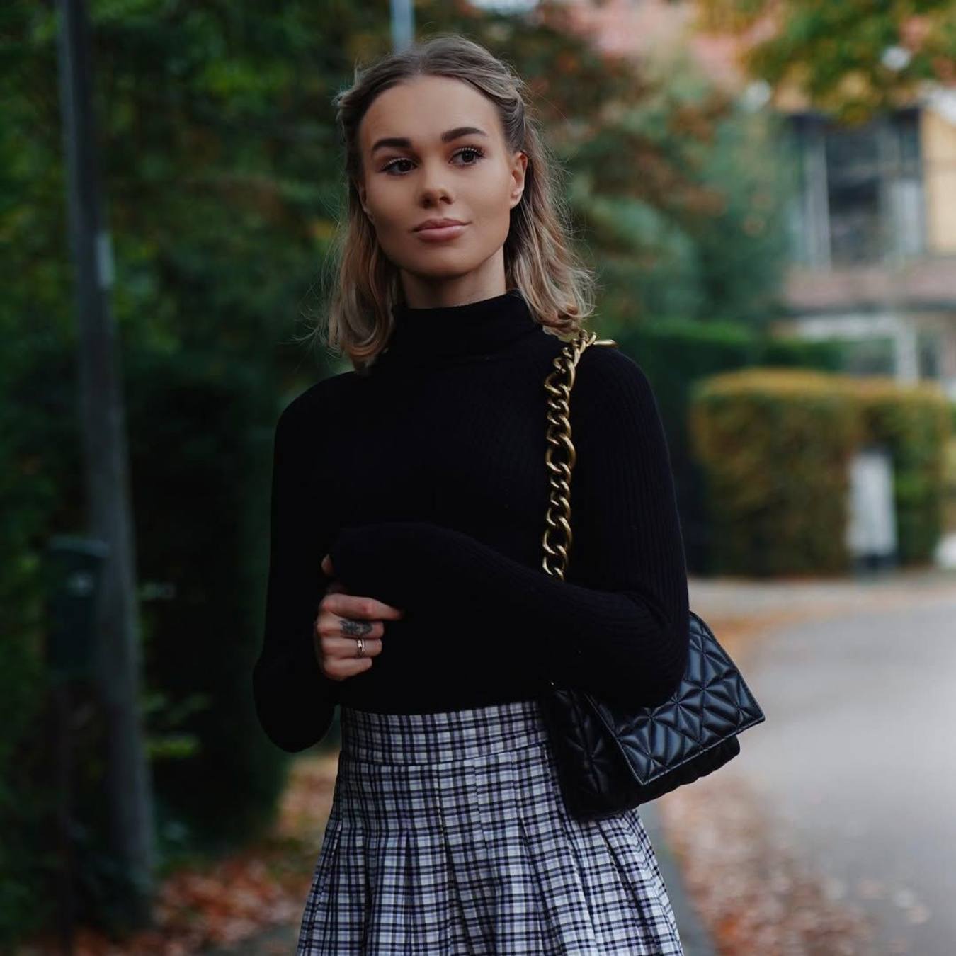 Een persoon met lichtbruin haar draagt ​​een zwarte coltrui en een grijze geruite plooirok, en heeft een zwarte tas met kettingriem bij zich.