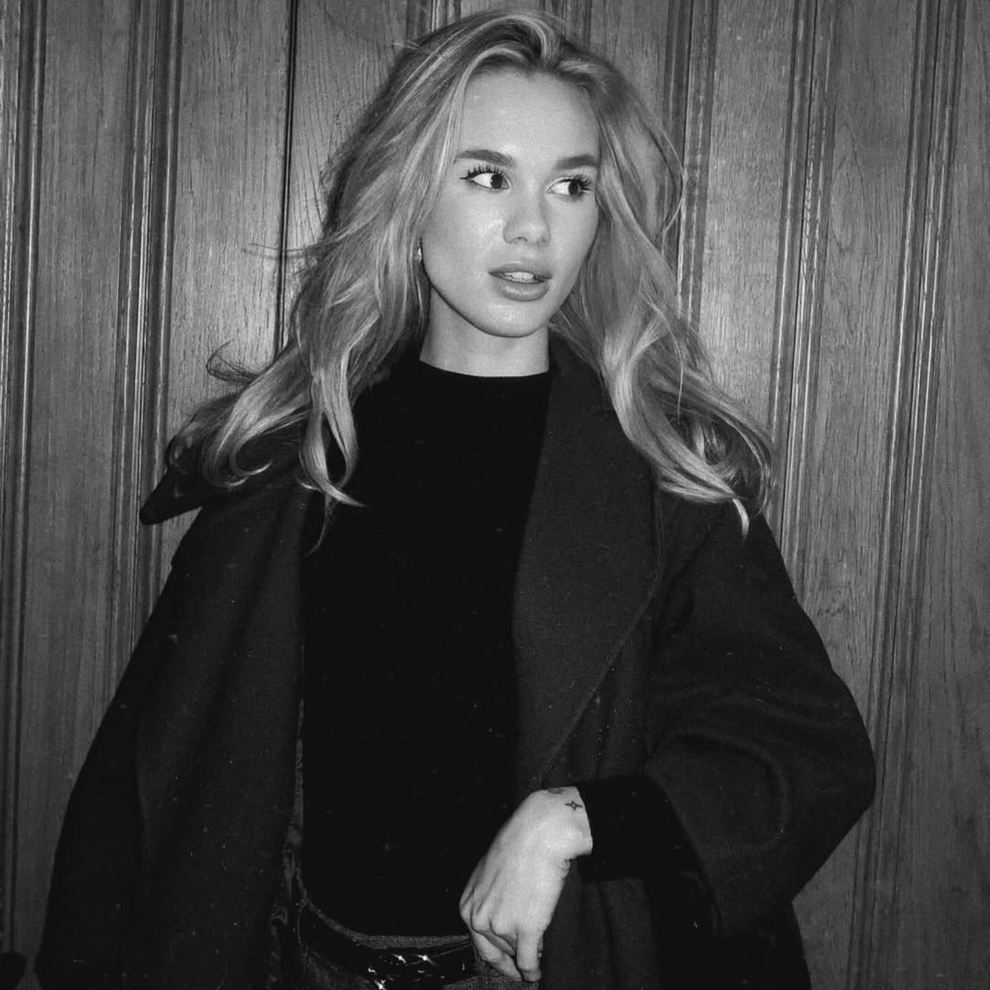 Een persoon met golvend haar, gekleed in een jas over een donker shirt, staat voor een zwart-wit houten lambrisering.