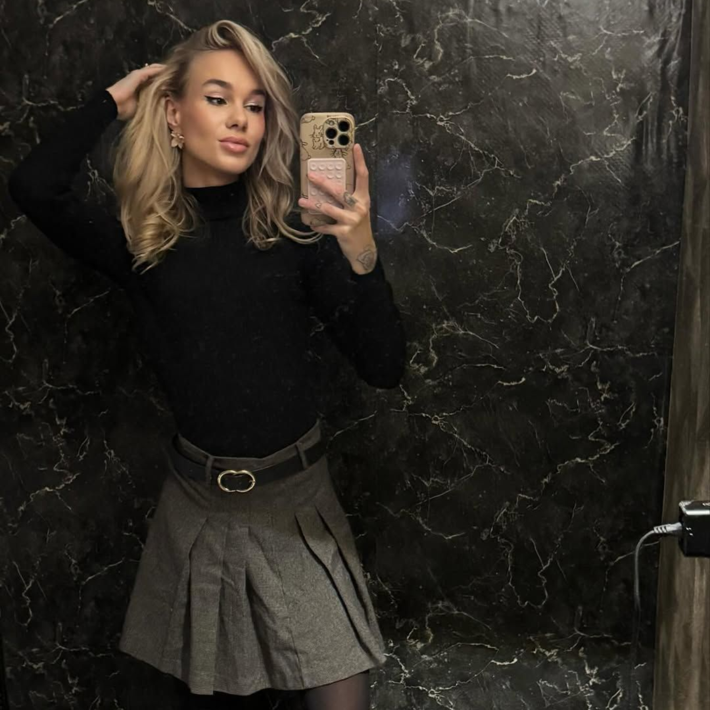 Een persoon met blond haar poseert voor een spiegel, gekleed in een zwarte coltrui, een grijze plooirok en een panty.