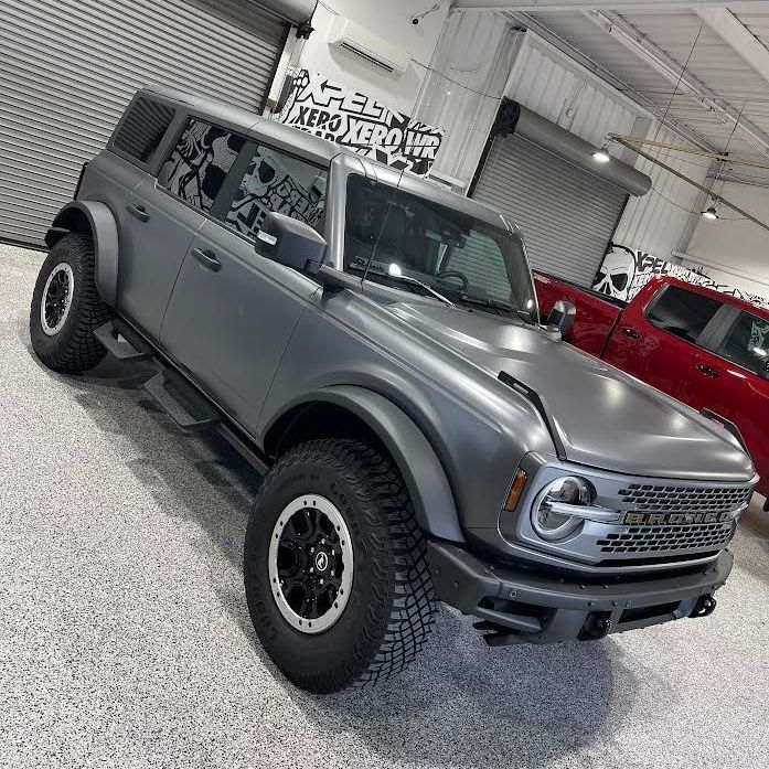 A gray ford bronco is parked in a garage next to a red truck.