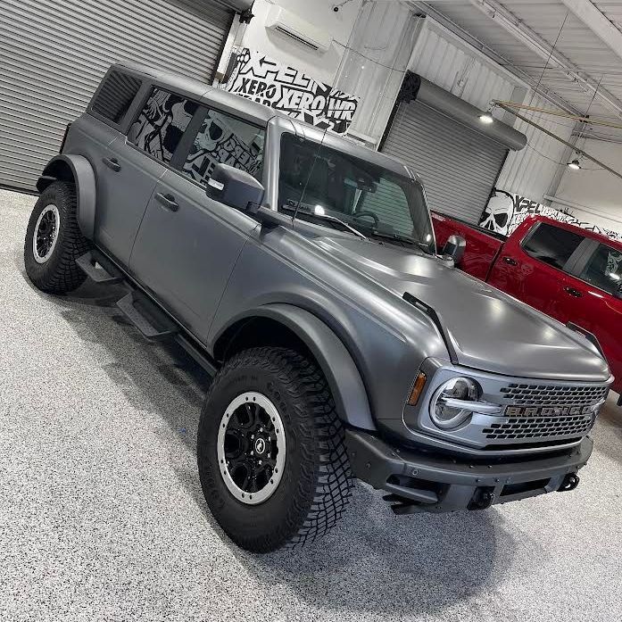 A gray ford bronco is parked in a garage next to a red truck.