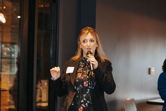 A person wearing a dark blazer over a floral blouse speaks into a microphone while gesturing in a modern indoor setting.