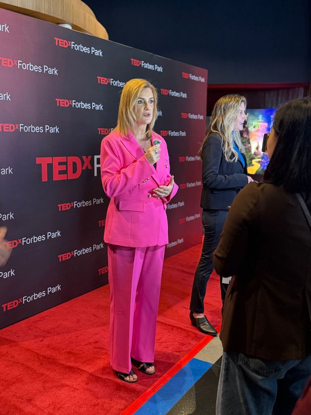A person in a bright pink suit speaks into a microphone on a red carpet in front of a TEDx Forbes Park branded backdrop.