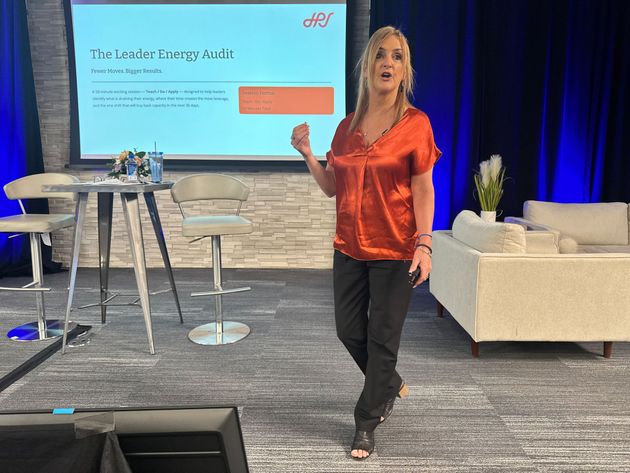 A speaker in a shiny orange top gestures toward a projected presentation slide while standing on a stage.