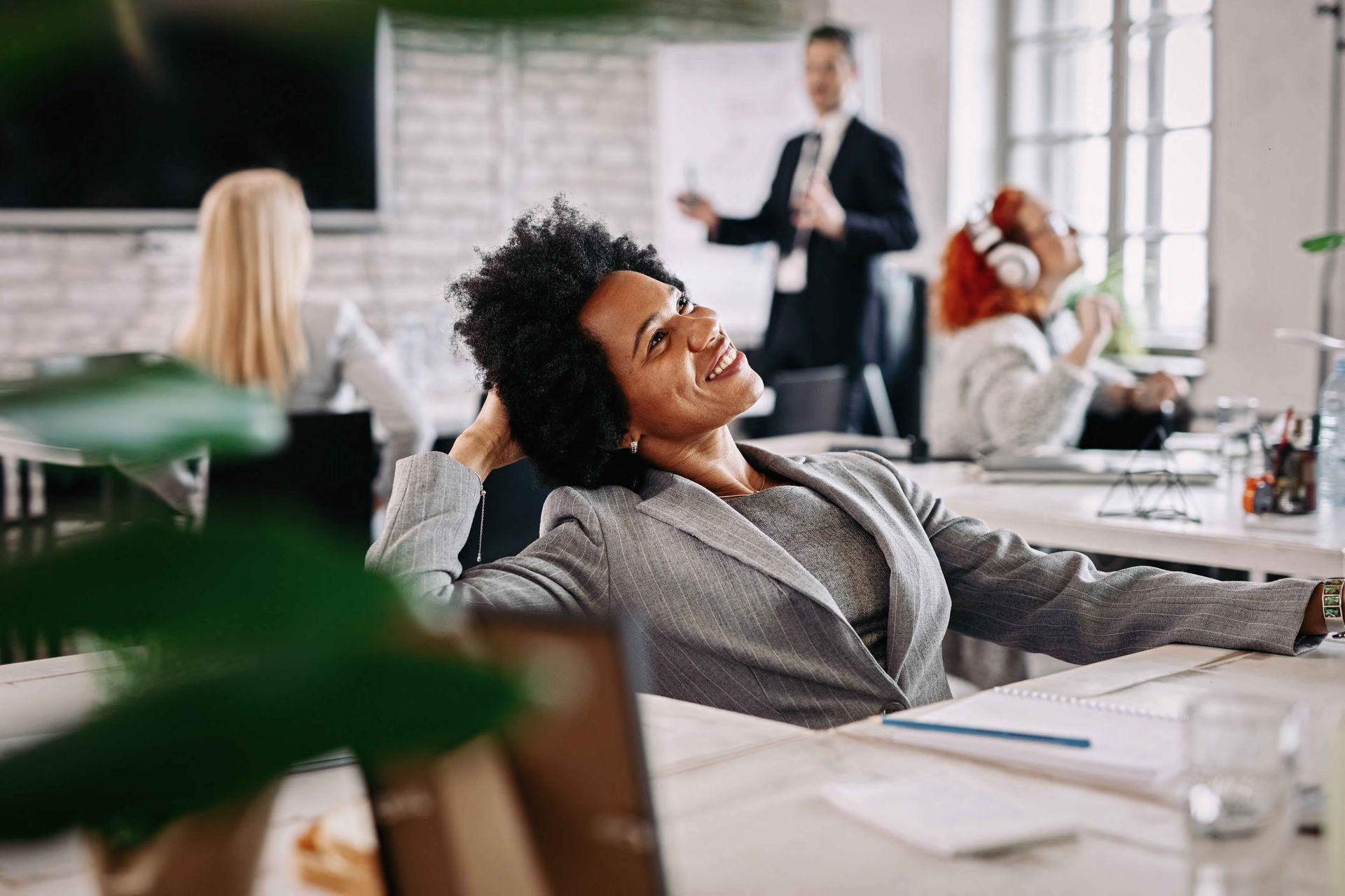 Smiling Black woman relaxing at her desk during a meeting, other colleagues in the background at a modern office.
