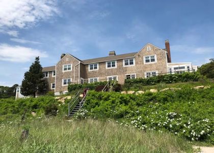 A large, shingled, multi-story house sits atop a grassy, flower-covered hill under a bright blue sky.