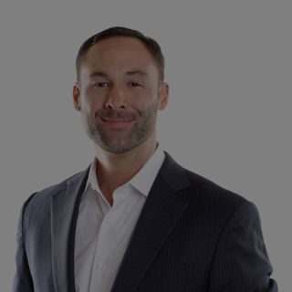 A man with a trimmed beard wearing a blazer and white shirt smiles slightly.