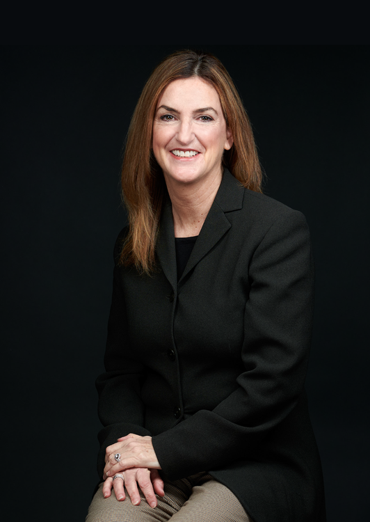 Woman in dark blazer smiles, seated against a dark background.