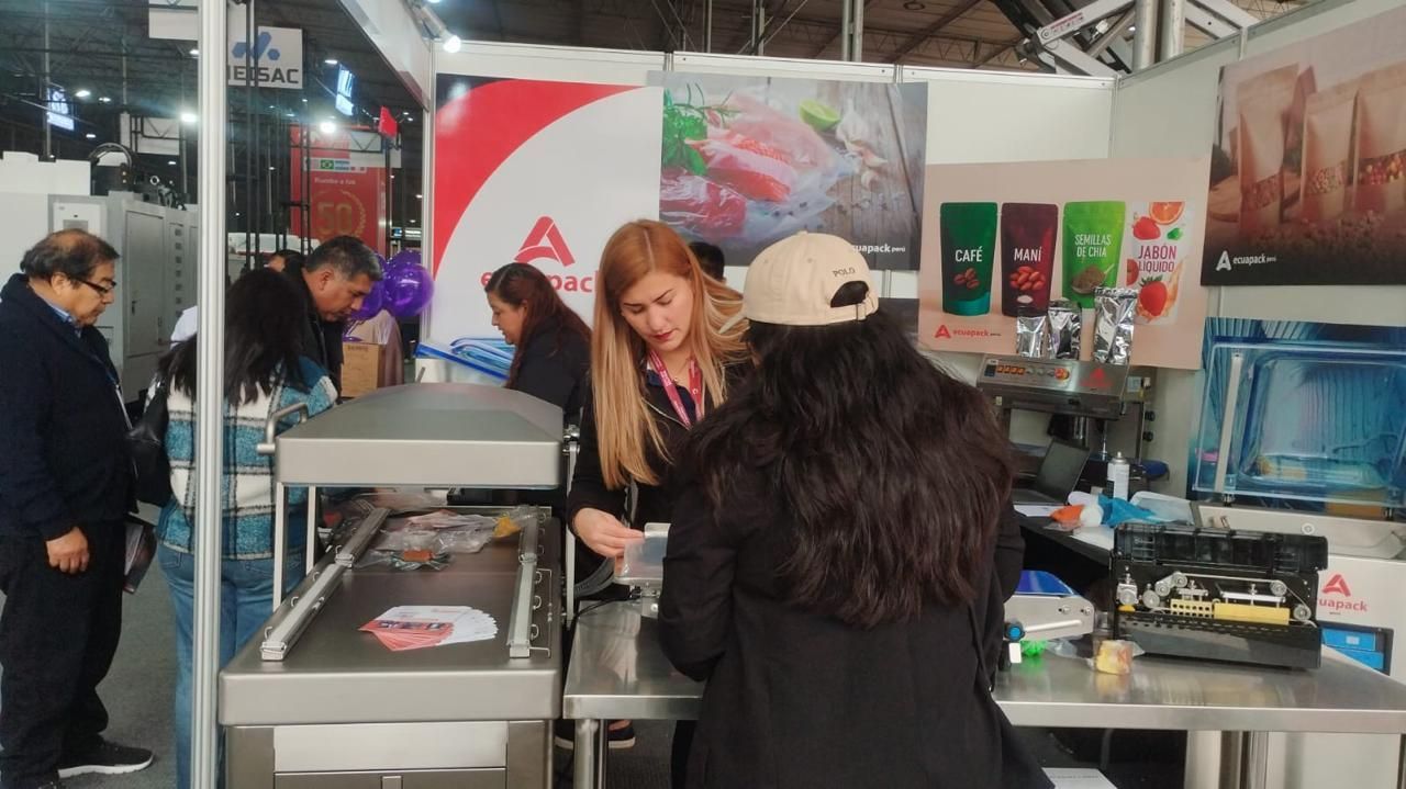 Personas en un stand de una feria comercial, mirando una pantalla de televisión; varios colores y artículos.
