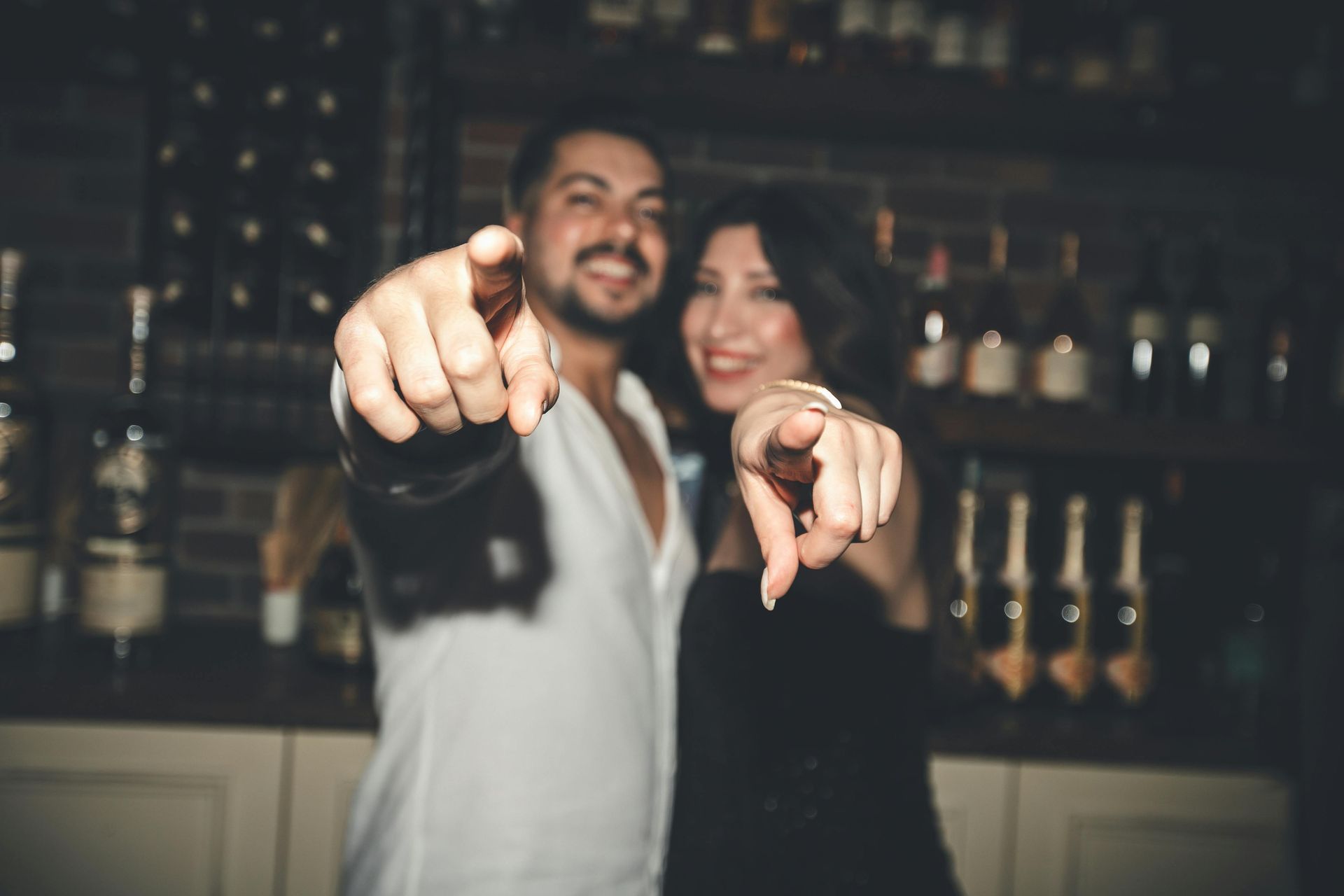 A man and a woman are pointing at the camera in a bar.