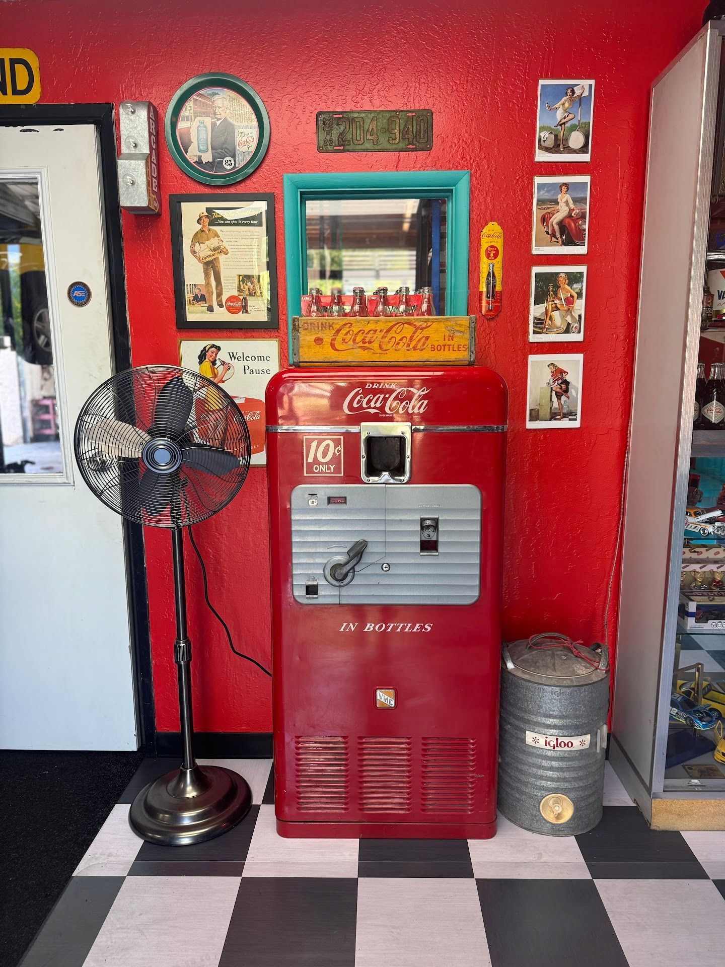 Vintage Red Coca-Cola Vending Machine with Memorabilia on a Red Wall | Greasers Automotive