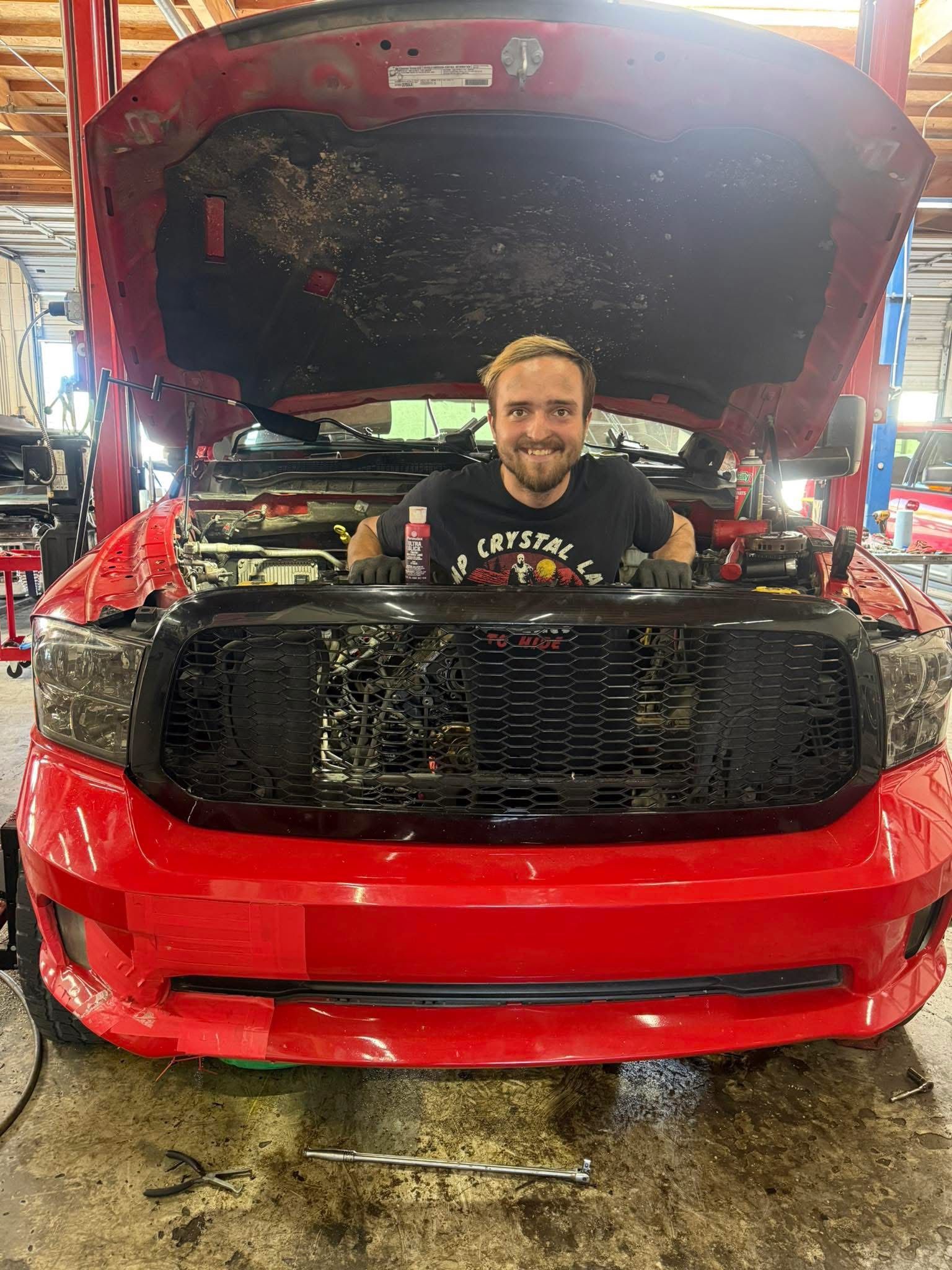 Man in black shirt smiles, standing inside the open hood of a red truck in a garage | Greasers Automotive